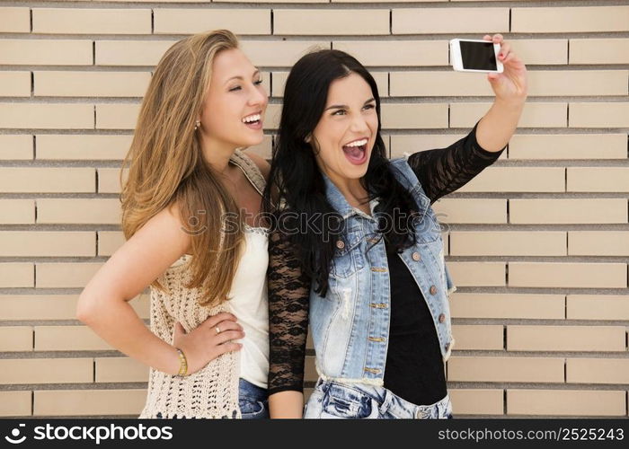 Two beautiful and young girlfriends taking pictures, in front of a brick wall