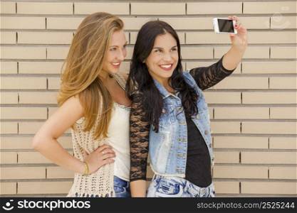 Two beautiful and young girlfriends taking pictures, in front of a brick wall