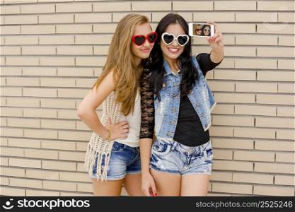 Two beautiful and young girlfriends taking pictures, in front of a brick wall