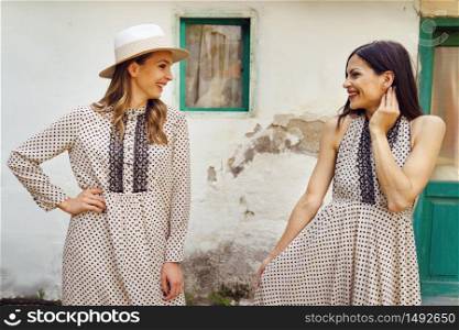 Two beautiful caucasian women standing in front of the old house wall wearing dress smiling and looking at each other talking in summer or spring day real people friendship and love concept