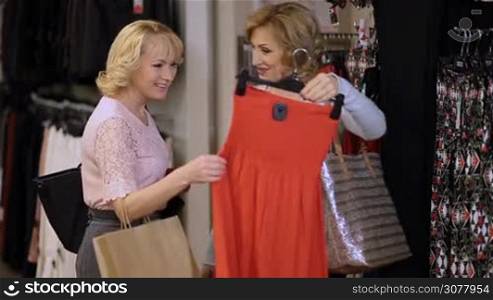 Two beautiful elderly women shopping in fashionable store together. Mature female selecting a dress and asking opinion of her cheerful friend while shopping for clothes in clothing shop.