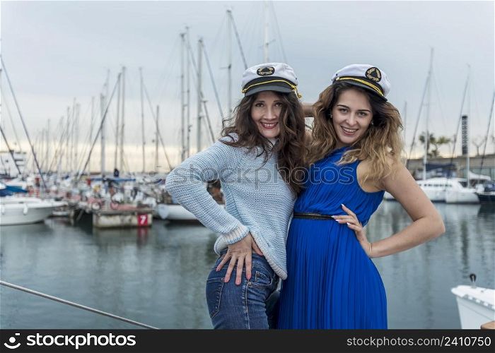Two beautiful model women standing and posing on a sailboat while looking at camera smiling
