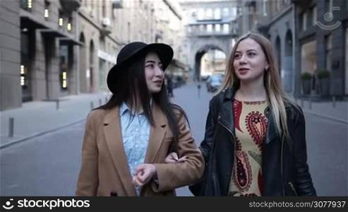 Two beautiful smiling women walking down the street on sunny day after good shopping. Stylish asian girl in hat and her attractive caucasian friend holding hands strolling city street in trade centre and chatting about latest gossips. Slow motion.