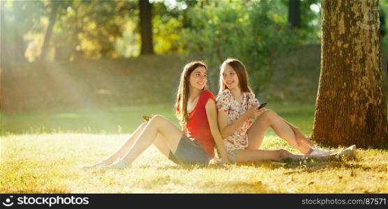 Two Beautiful Teenagers Holding Mobil Phones Sitting under a Tree in the Bright Summer Day