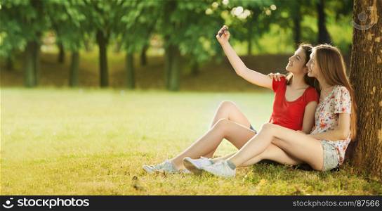 Two Beautiful Teenagers Make Selfies, Sitting under a Tree in the Bright Summer Day