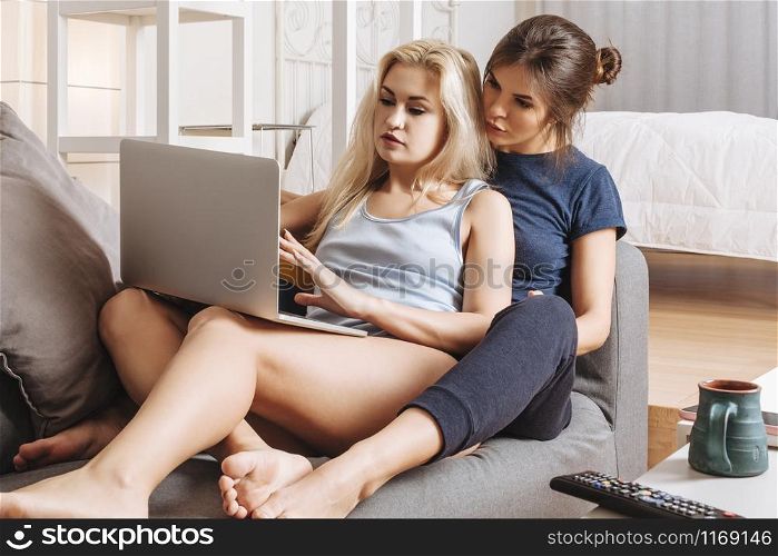 Two beautiful women in a relationship sitting on a sofa using a laptop together.