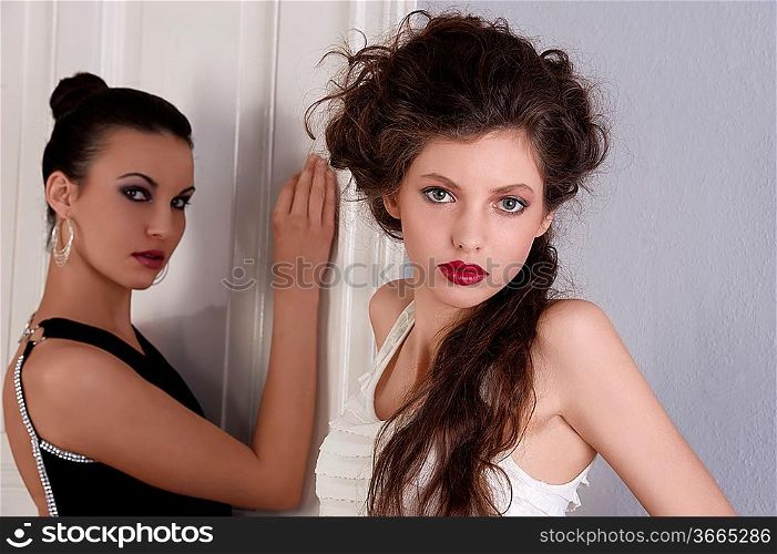 two beautiful women with hair style and elegant dress posing indoor near an old fashion door looking towards the camera