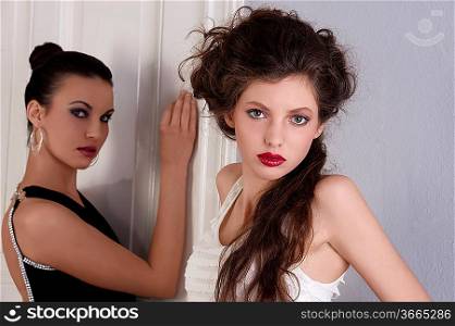 two beautiful women with hair style and elegant dress posing indoor near an old fashion door looking towards the camera