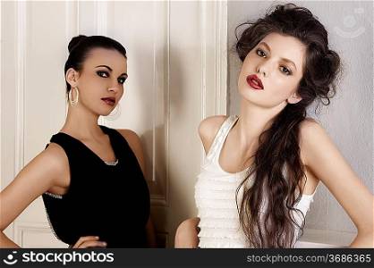two beautiful women with hair style and elegant dress posing indoor near an old fashion door posing while looking towards camera