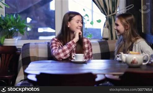 Two beautiful young girlfriends sitting in modern coffee shop and talking with happy smiles while waiting for their friends. Cute teenage girl arrives and joins her female friends in cafe for a chat. Group of girl friends meeting in cafe