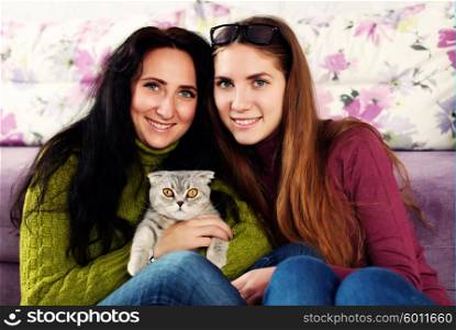 Two beautiful young sisters playing with British breed pet kitten at home. Indoors, lifestyle