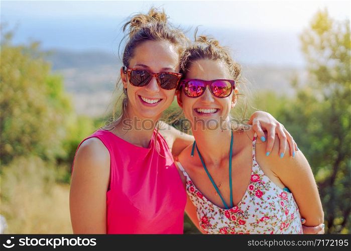 Two beautiful young woman female girl friends or sisters having fun in nature outdoors hugging embracing each other smiling wearing summer dress sunglasses in the autumn travel