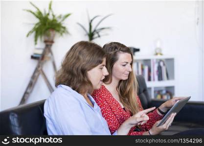 Two beautiful young women at home sitting on sofa or settee using a tablet PC computer and smiling