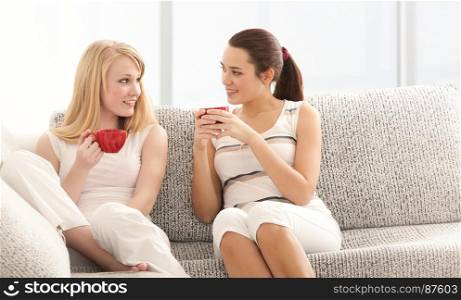 Two Beautiful Young Women Drinking Coffee, Talking to each Other, Sitting on the Sofa in Living Room