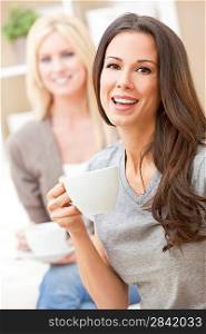 Two beautiful young women friends or girls with perfect smiles drinking tea or coffee from a white cup at home on a sofa