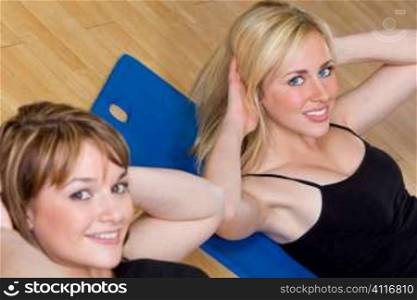 Two beautiful young women, one blond, one brunette, working out on on mats doing aerobic sit ups at the gym. The focus is on the blond girl with blue eyes.