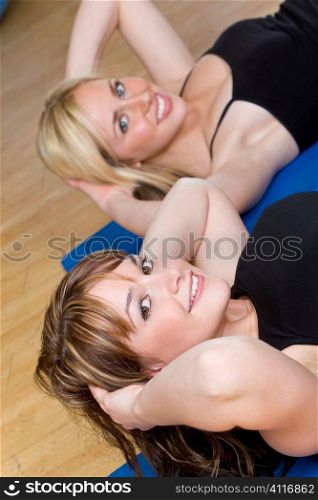 Two beautiful young women, one blond, one brunette, working out on on mats doing aerobic sit ups at the gym. The focus is on the brunette girl with brown eyes.