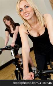 Two beautiful young women, one blond, one brunette, working out on spinning exercise bikes at the gym