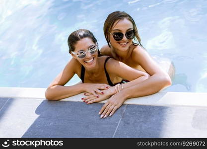 Two beautiful young Women with sunglasses relaxing together by poolside of outdoor swimming pool and smiling