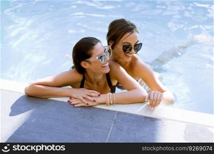Two beautiful young Women with sunglasses relaxing together by poolside of outdoor swimming pool and smiling