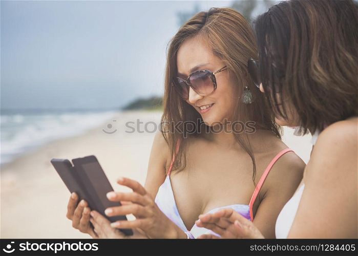 two beautiful younger woman wearing bikini looking on smart phone beside vacation sea beach