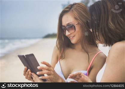 two beautiful younger woman wearing bikini looking on smart phone beside vacation sea beach