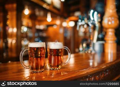 Two beer mugs on wooden bar counter, macro, nobody. Octoberfest symbol or concept. Glasses with golden beverage and foam on the table in pub, closeup view, blur background