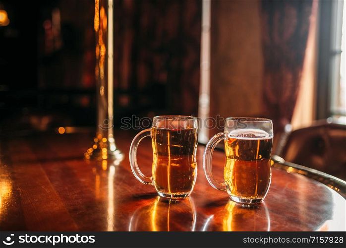 Two beer mugs on wooden bar counter, macro, nobody. Octoberfest symbol or concept. Glasses with golden beverage and foam on the table in pub, closeup view, blur background. Two beer mugs on wooden bar counter, nobody