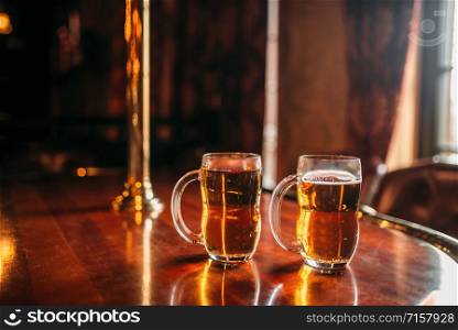 Two beer mugs on wooden bar counter, macro, nobody. Octoberfest symbol or concept. Glasses with golden beverage and foam on the table in pub, closeup view, blur background. Two beer mugs on wooden bar counter, nobody