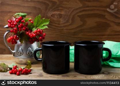 Two black campfire enamel coffee mug mockup with antique jug and cranberry branches. Empty mug mock up for design promotion.