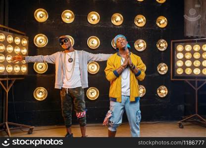 Two black rappers in caps, artists poses on stage with spotlights on background. Rap performers on scene with lights, underground music. Two black rappers in caps, artists poses on stage
