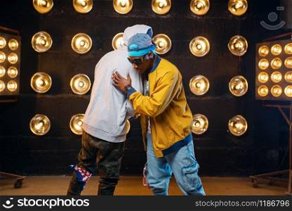 Two black rappers in caps hugging on stage, perfomance in club with spotlights on background. Rap performers on scene with lights, underground music concert, urban style