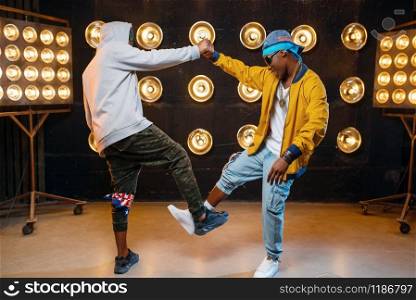 Two black rappers in caps hugging on stage, perfomance in club with spotlights on background. Rap performers on scene with lights, underground music concert, urban style. Two black rappers in caps hugging on stage