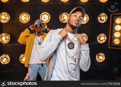 Two black rappers in caps on stage with spotlights on background. Rap performers on scene with lights, underground music, urban style. Two black rappers in caps on stage with spotlights