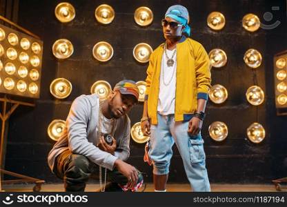 Two black rappers in caps, perfomance in club, stage with spotlights on background. Rap performers on scene with lights, underground music concert, urban style