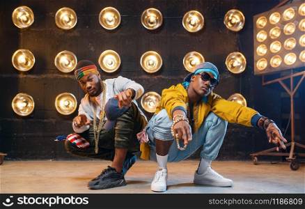 Two black rappers sitting on the floor, perfomance on stage with spotlights on background. Rap performers on scene with lights, underground music, urban style. Two black rappers sitting on the floor