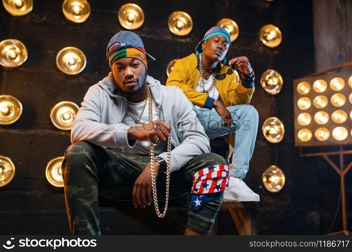 Two black rappers sitting on the steps, perfomance on stage with spotlights on background. Rap performers on scene with lights, underground music, urban style. Two black rappers sitting on the steps