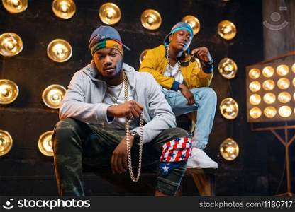 Two black rappers sitting on the steps, perfomance on stage with spotlights on background. Rap performers on scene with lights, underground music, urban style. Two black rappers sitting on the steps