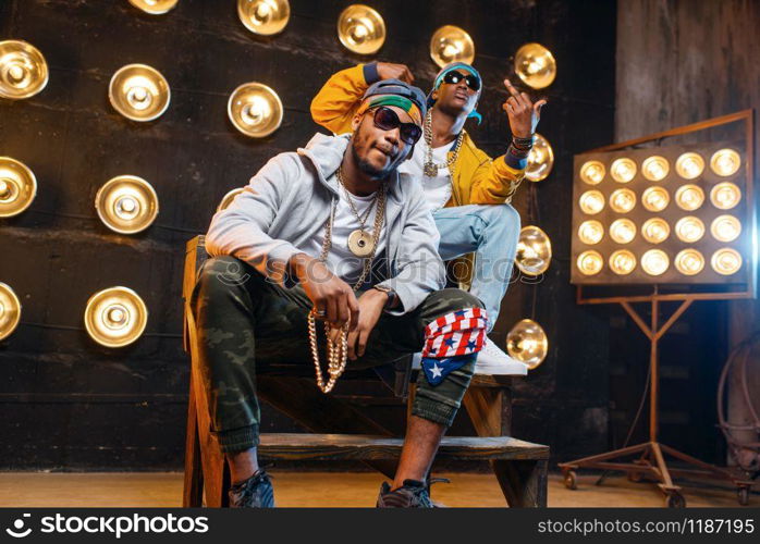 Two black rappers sitting on the steps, perfomance on stage with spotlights on background. Rap performers on scene with lights, underground music, urban style. Two black rappers sitting on the steps