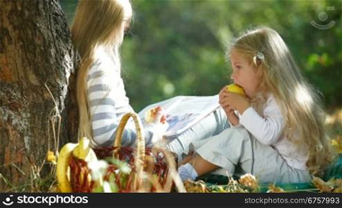 Two blond little girls under tree enjoying sunny autumn day, reading book and eating an apple
