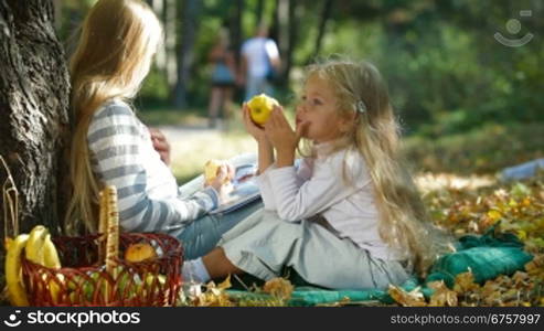 Two blond little girls under tree enjoying sunny autumn day, reading book and eating an apple