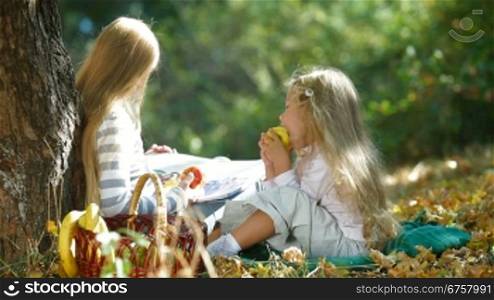 Two blond little girls under tree enjoying sunny day, reading book and eating an apple