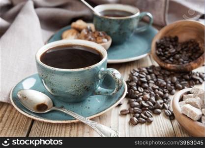 Two blue cups of black coffee, sugar cubes in a wooden bowls and scattered coffee beans on a background of a linen cloth