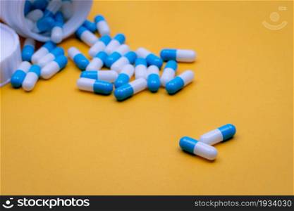 Two blue-white capsules pills on blur many of pills pouring from plastic drug bottle. Capsule pills on yellow background. Prescription drugs. Pharmaceutical industry. Health care and medicine concept.