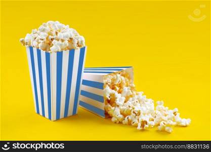 Two blue white striped carton buckets with tasty cheese popcorn, isolated on yellow background. Box with scattering of popcorn grains. Movies, cinema and entertainment concept.