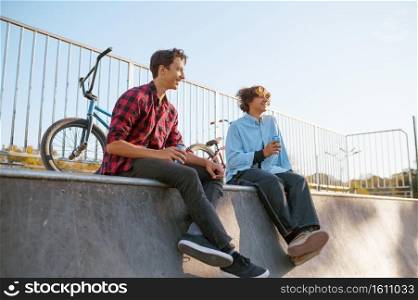 Two bmx bikers leisures in skatepark after training. Extreme bicycle sport, dangerous cycle exercise, street riding, biking in summer park. Bmx bikers leisures in skatepark after training