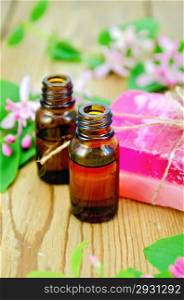 Two bottles of aromatic oil, pink homemade soap, branches with leaves and pink flowers of honeysuckle on a background of wooden boards
