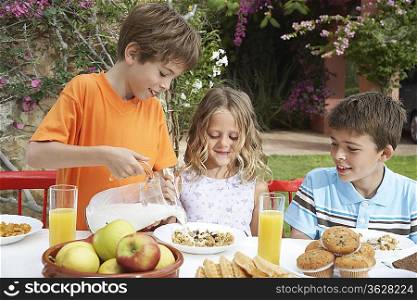 Two boys and girl (6-11) at table, boy pouring milk on cereal, garden