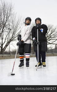 Two boys in ice hockey uniforms holding hockey sticks standing on ice rink in ice skates.