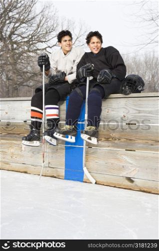 Two boys in ice hockey uniforms sitting on ice rink sidelines looking and smiling.
